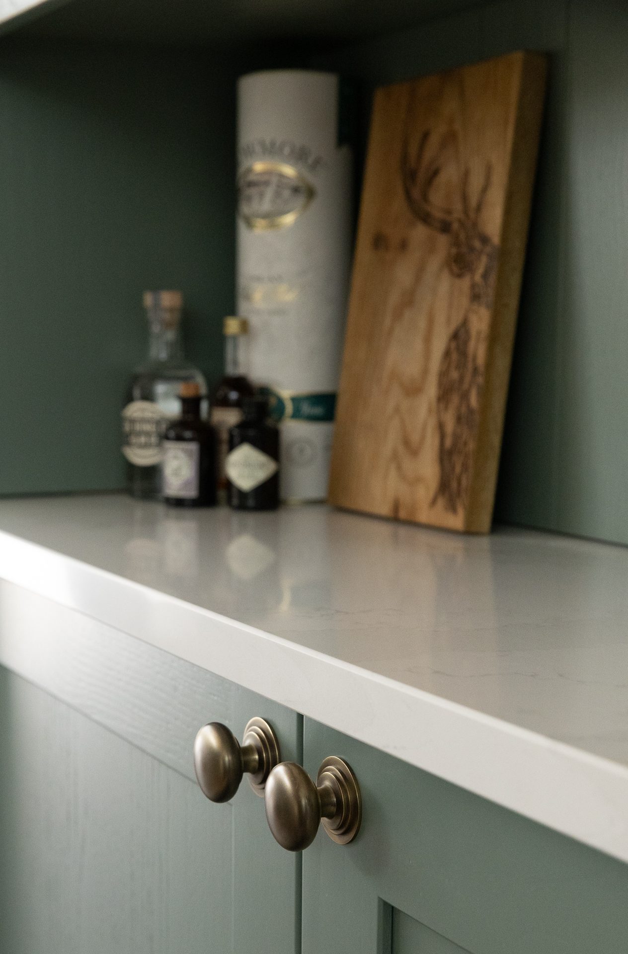 A sage green drinks cabinet with a pale stone worktop, a whisky tube, small apothecary bottles and a stag-etched wooden board, antique nickel knobs below, header image for the Brushed chapter