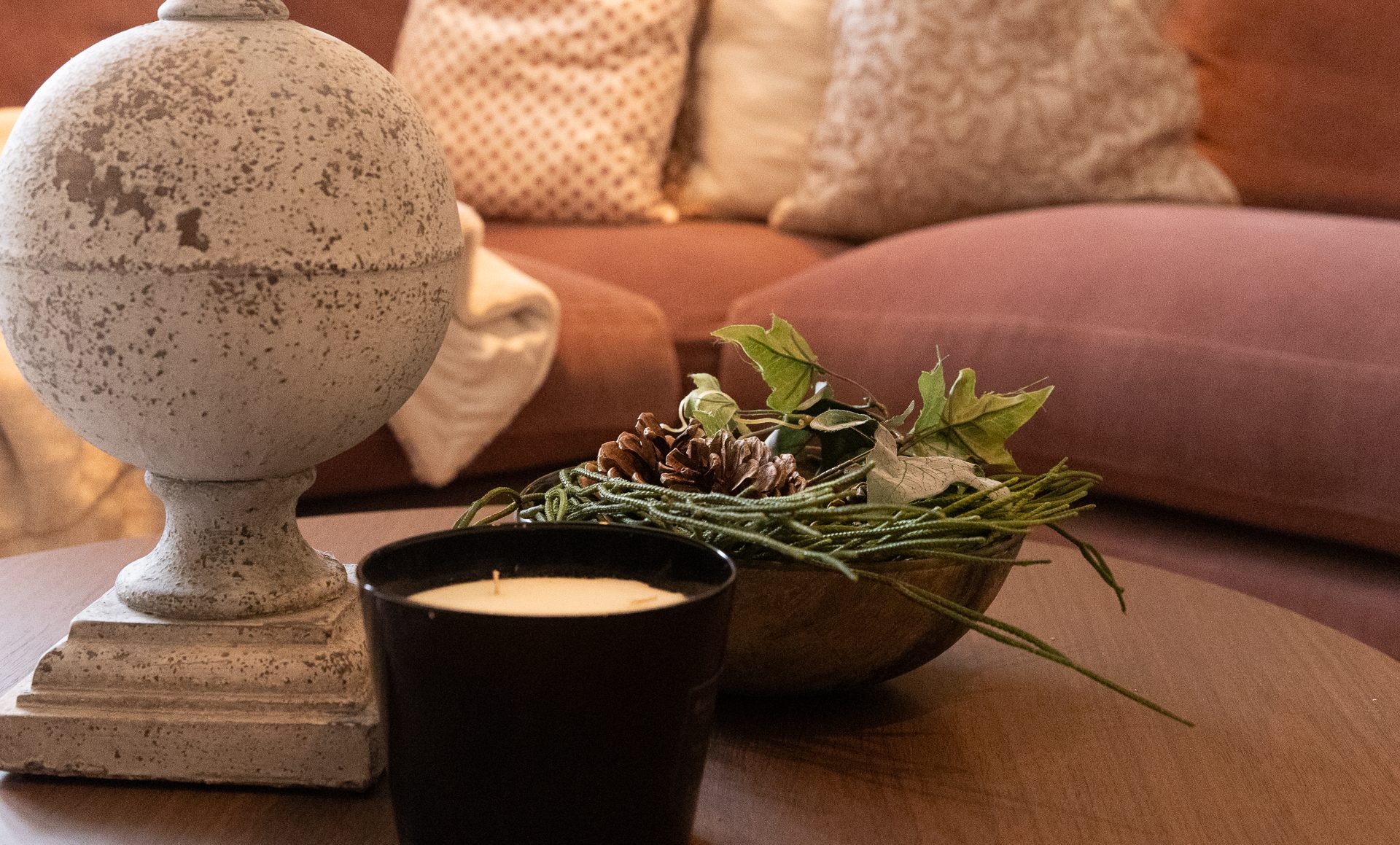 A weathered stone urn finial, a small unlit black candle and a wooden bowl of pinecones and ivy on a round wooden coffee table, a terracotta sofa with cream patterned cushions behind