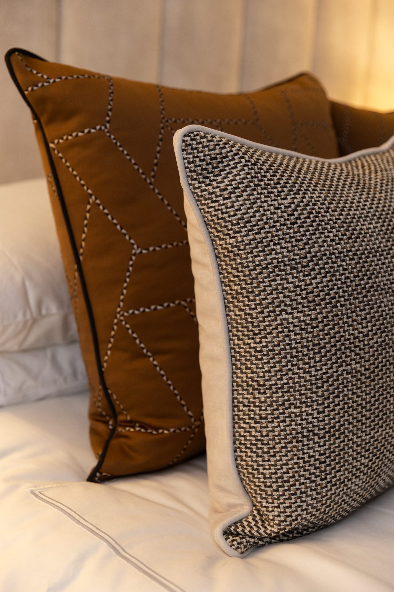 A tan leather-effect cushion with stitched geometric lines and a greige tweed cushion with cream piping, layered on white bedlinen against a taupe panelled headboard, with a warm lamp to the right