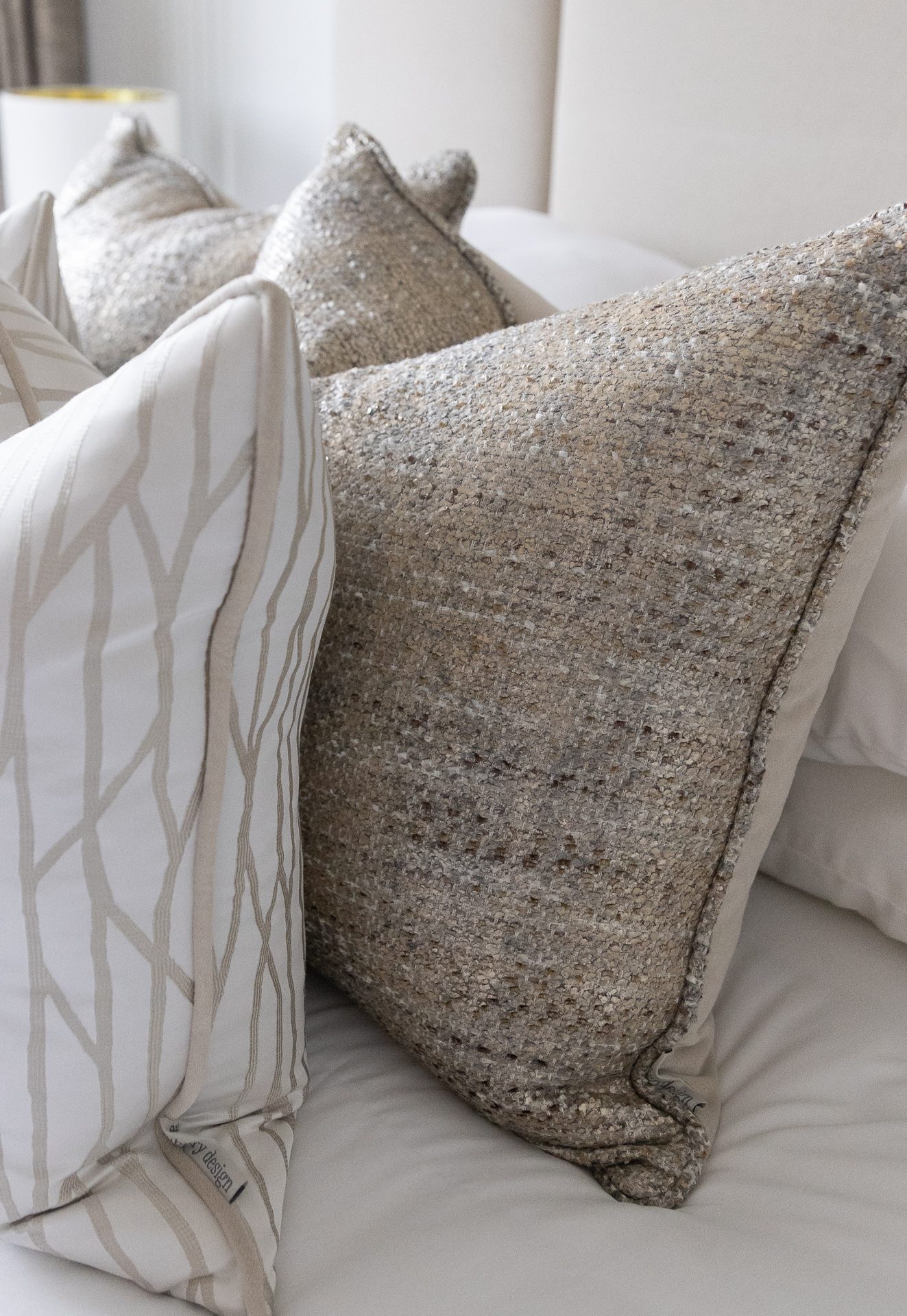 A cream cushion with a fine pale-linear pattern in front of a larger greige bouclé cushion, on white bedlinen with a table lamp behind