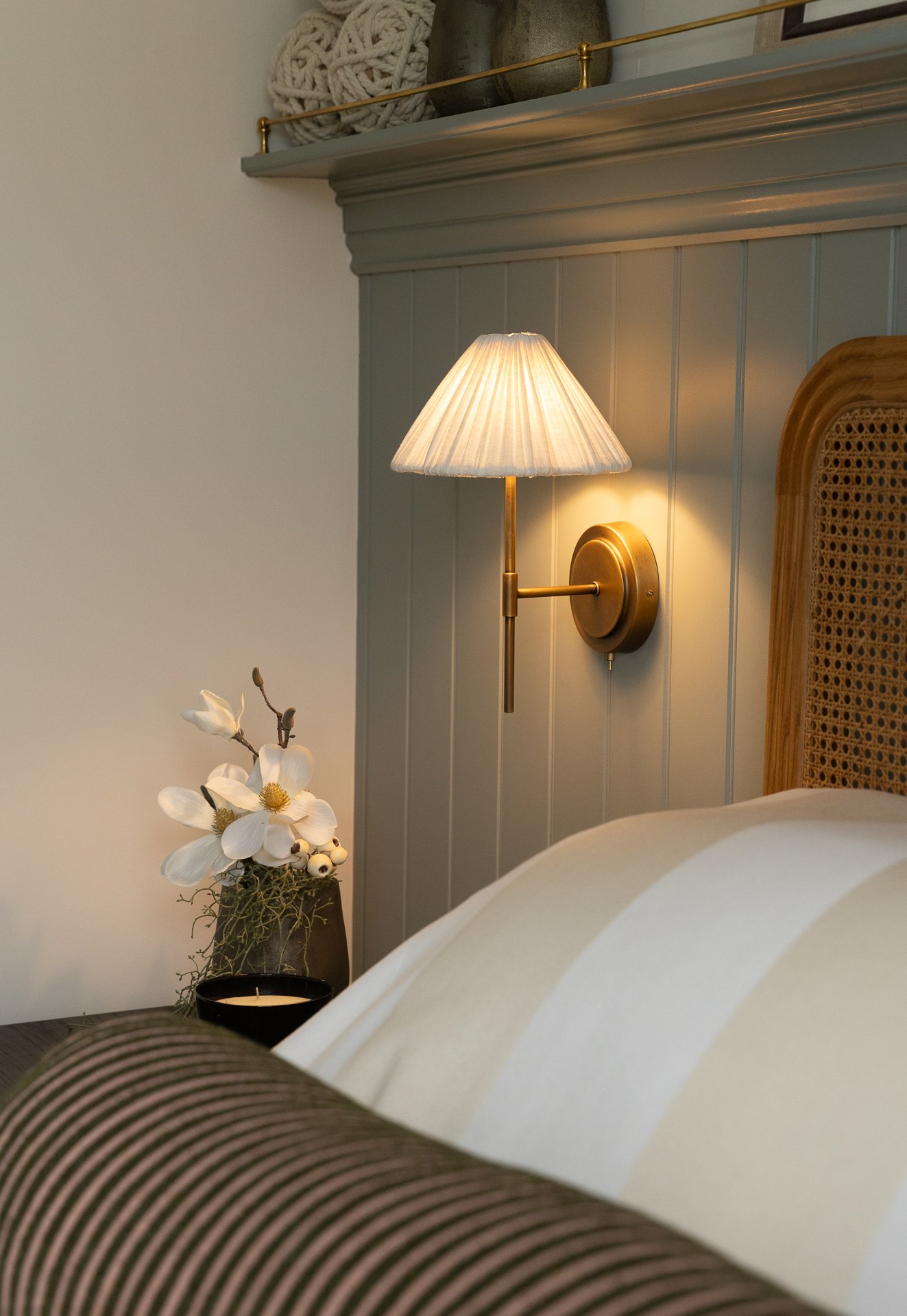 Brass wall sconce with a pleated cream shade beside the bed, magnolia in a stone vessel and a black candle on the side