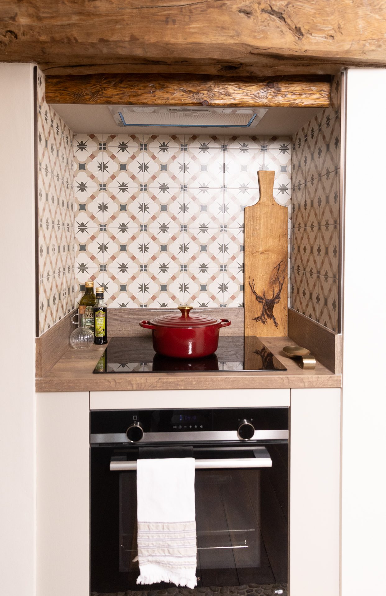 The Bamburgh range nook, a patterned tile backsplash held under a reclaimed oak beam, induction hob with a small red Le Creuset pot and a stag-etched oak serving board to the side