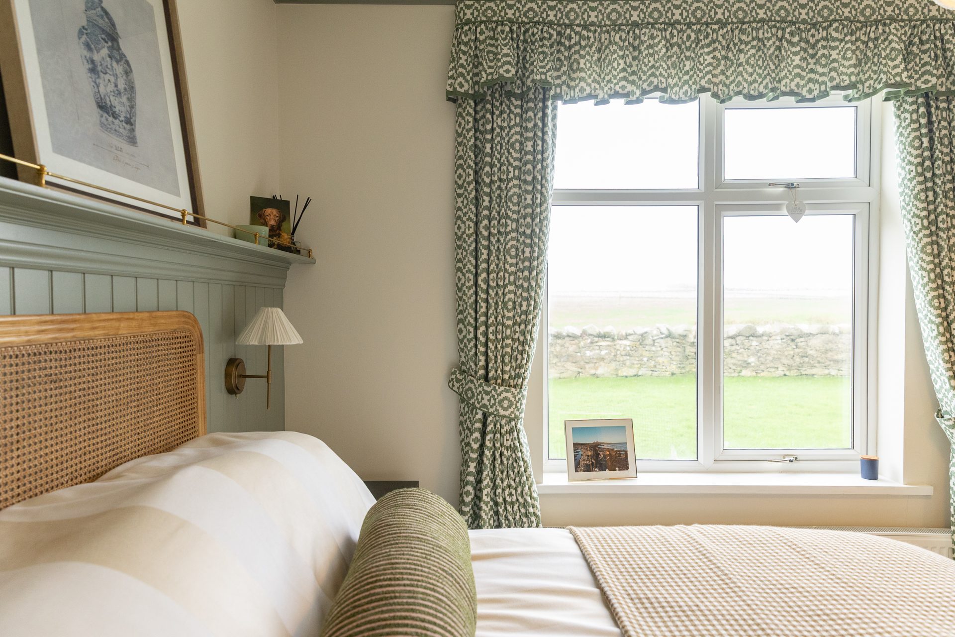 A Bamburgh guest bedroom, a rattan headboard against sage panelling, patterned curtains, a drystone wall and field visible through the window