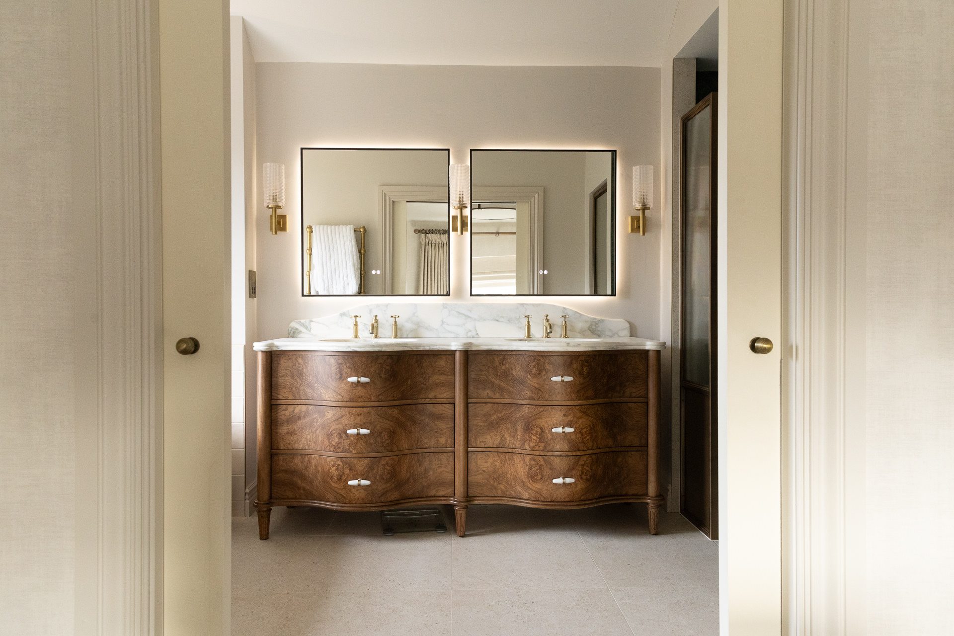 The en-suite vanity in burl wood with twin backlit mirrors, marble counter and brass fittings, seen through the open cabinetry doors