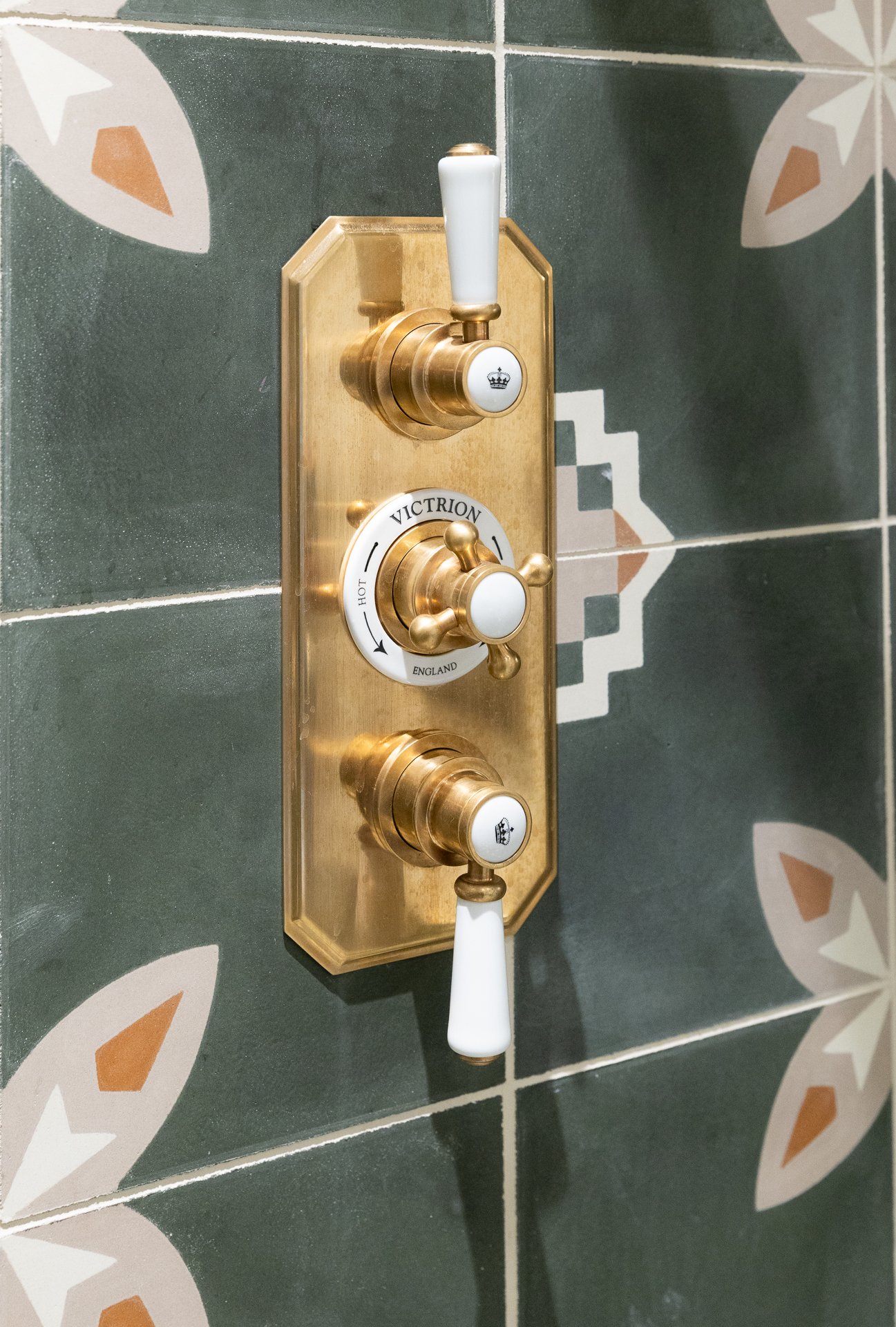 Close detail of a Victrion brass thermostatic shower valve with three white ceramic handles, set against the green encaustic tile