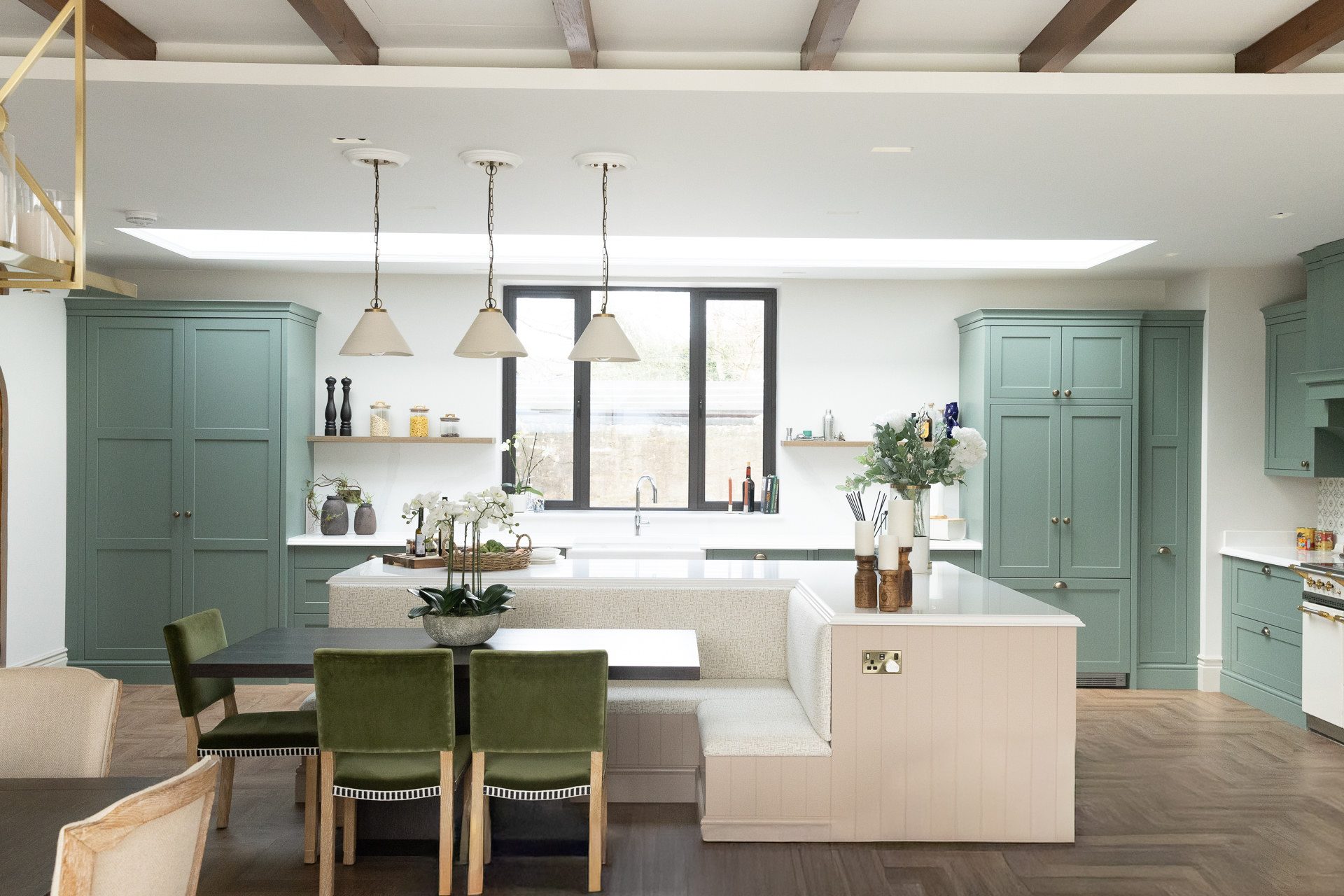 Wide view of the Elmfield Road sage green kitchen with three cream pendants over the island, a built-in upholstered banquette with green velvet chairs, oak beams and parquet floor