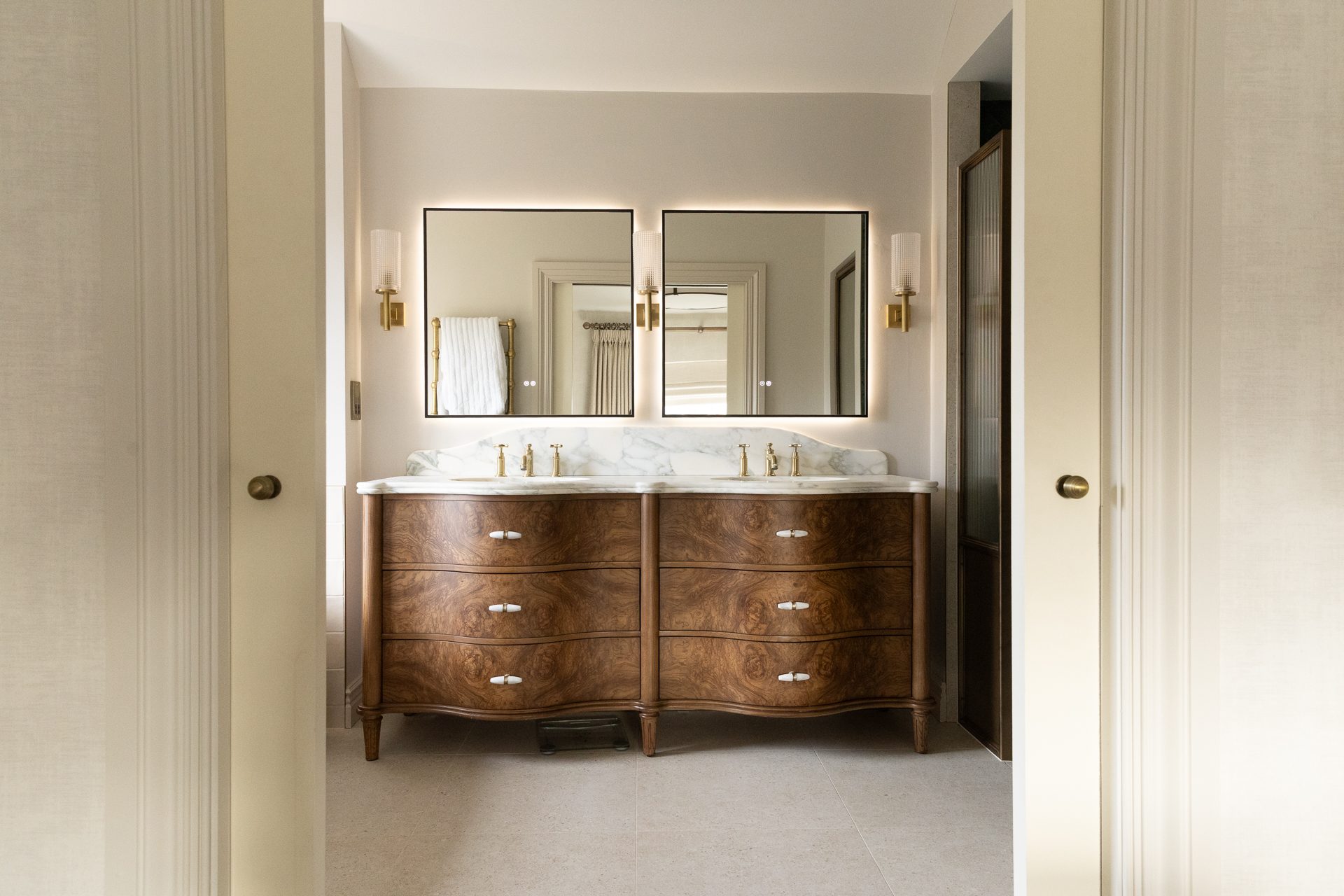 The master bathroom at Elmfield Road, viewed through the doorway, a walnut burl double vanity with twin brass taps, twin backlit mirrors and twin brass wall sconces