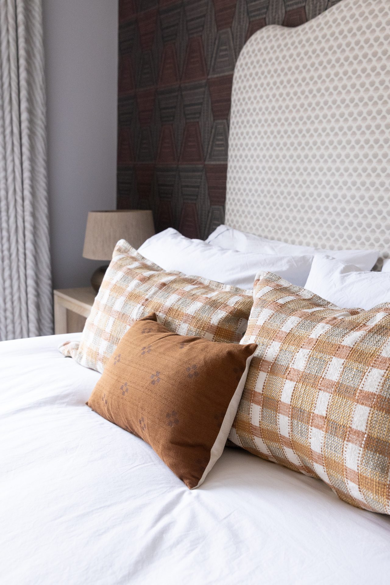 A guest bedroom with soft plaster walls, a patterned upholstered headboard, and layered linens in plaid and rust velvet