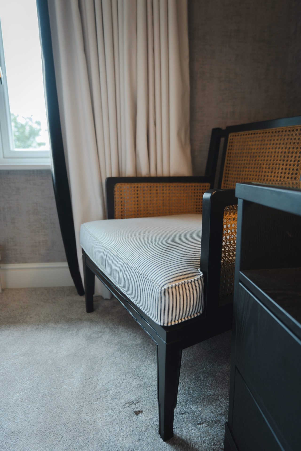A caned-back armchair with a black and cream striped seat, sitting by the bedroom window