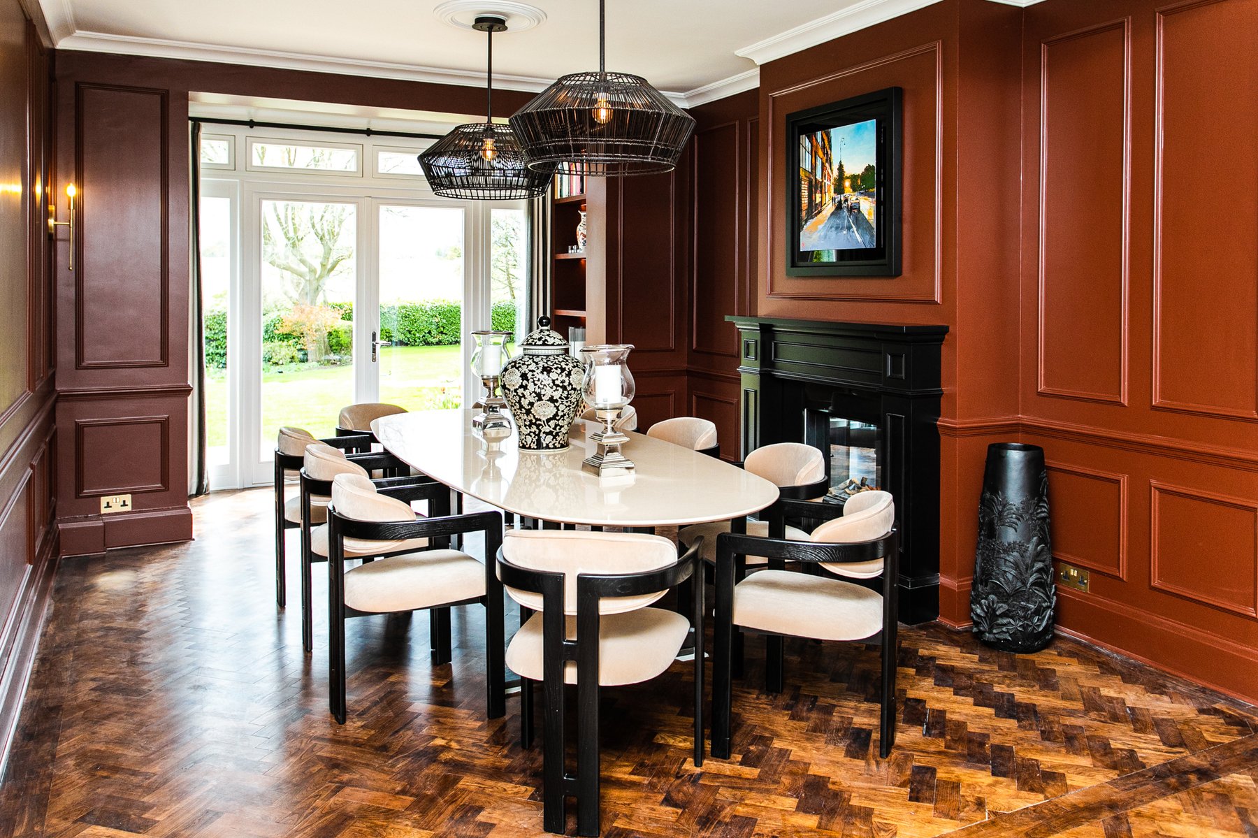 The Montagu dining room, deep terracotta-red panelled walls, oval table with cream chairs, a pair of woven black pendants overhead, herringbone parquet floor, french doors to the garden at the end of the room