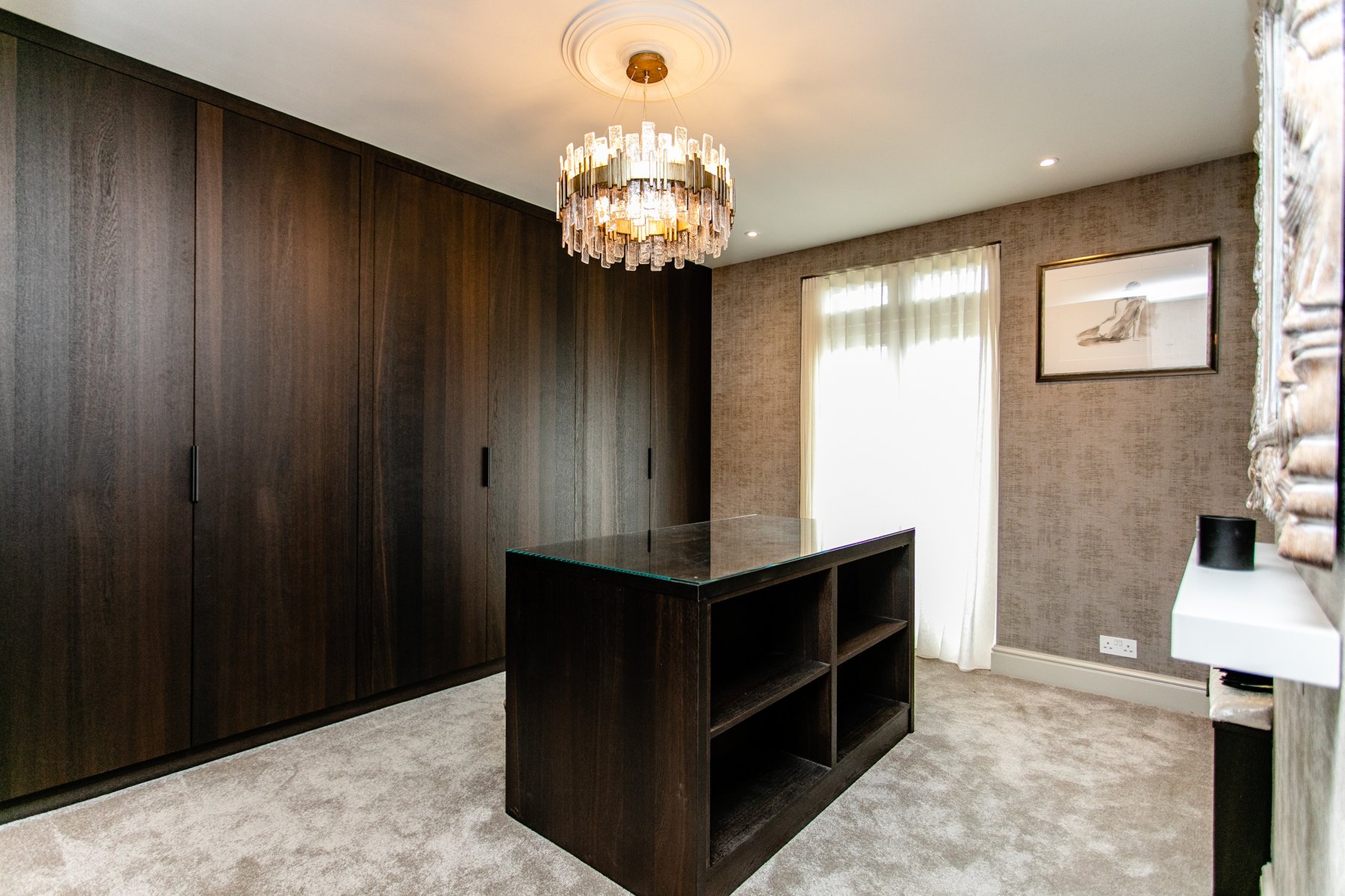 The Montagu dressing room in its architectural state, dark walnut wardrobe doors running the length of the walls, a central open-shelf island, crystal chandelier under a plaster ceiling rose, a cream silk drape to the window