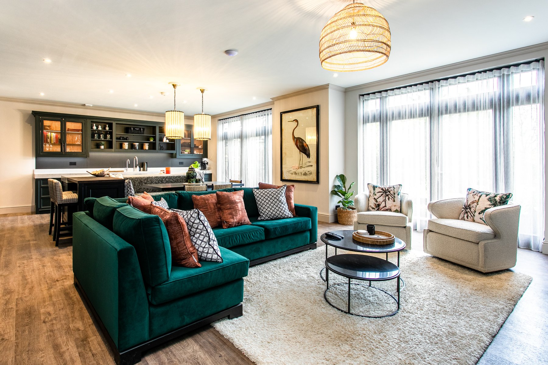 The Montagu kitchen and family room, a teal velvet sofa facing two cream boucle armchairs, a rattan pendant overhead, a framed heron print on the wall, the kitchen island visible in the background