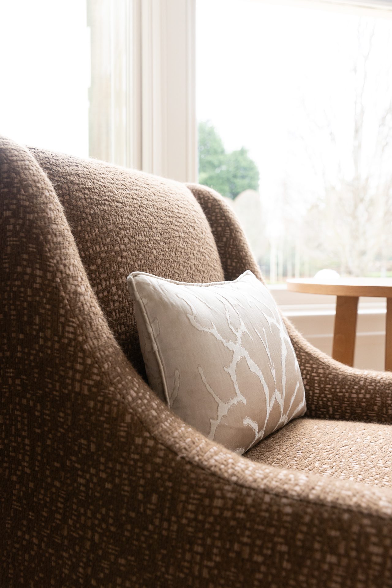 A brown tweed chair held in bay-window light, a single cream branches-patterned cushion set against it