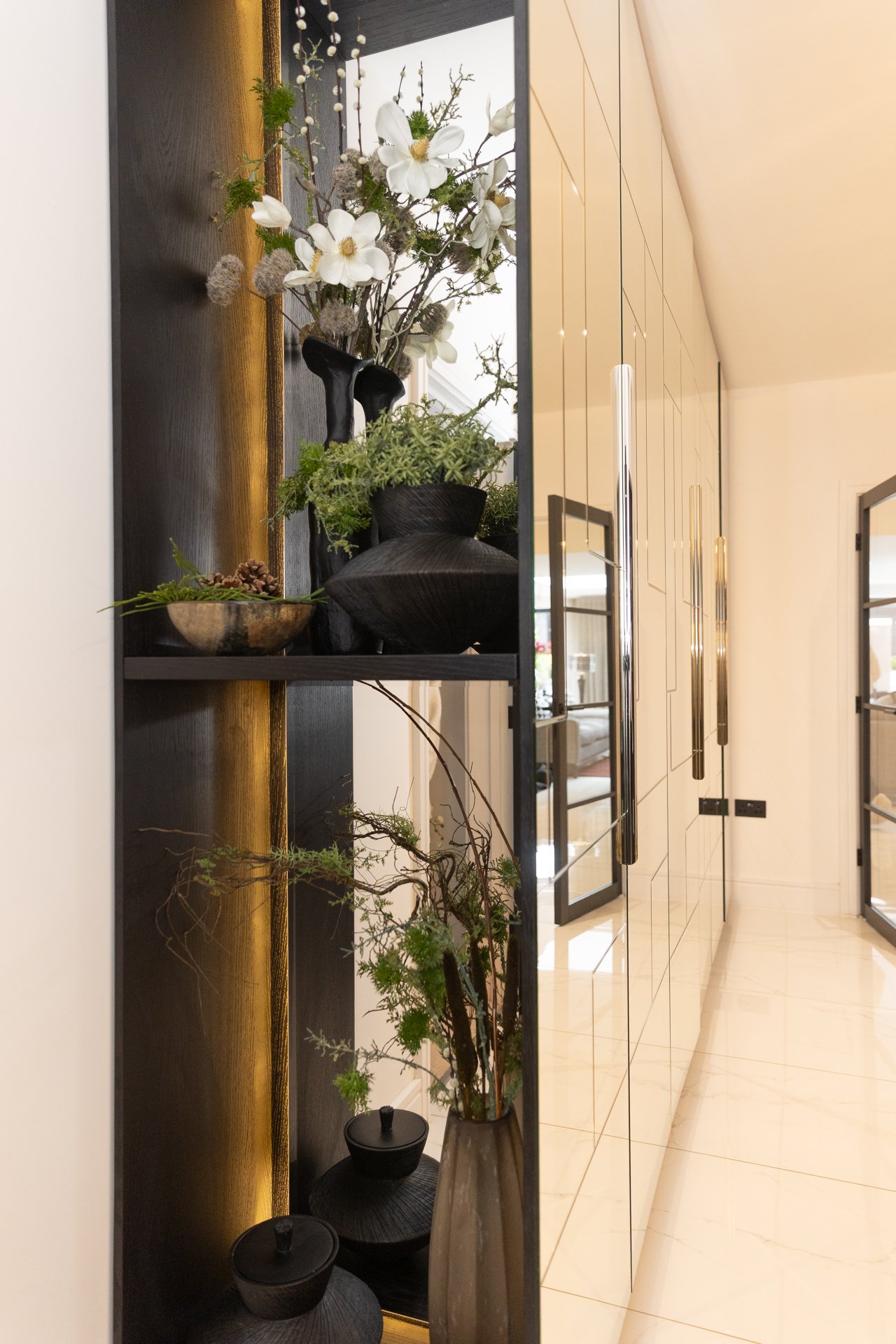 The Sycamore hallway, a dark oak shelf-niche with white magnolias in a black vase and lit from behind in amber, next to a cream panelled corridor with black-framed glass doors and long brass handles
