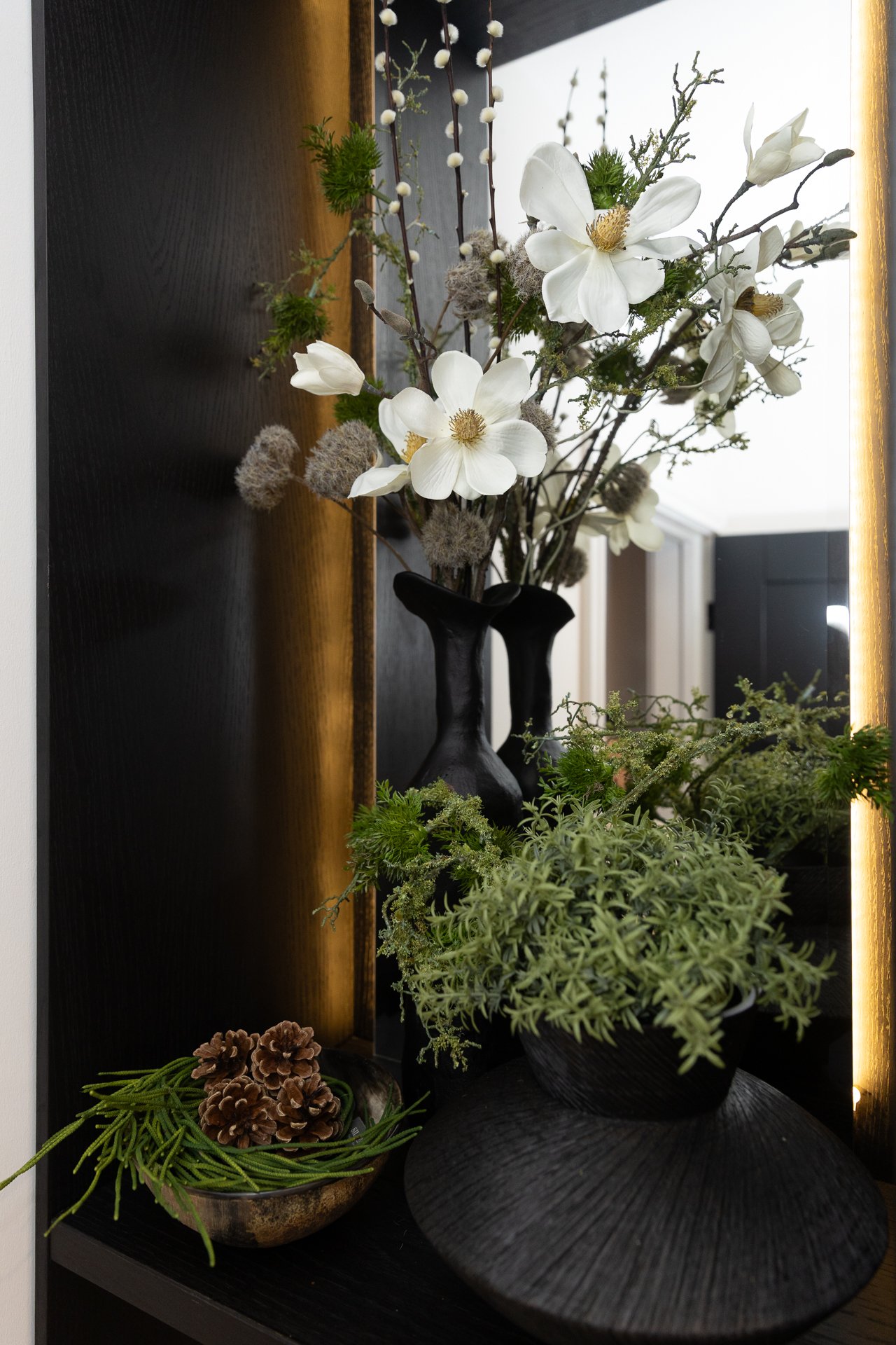 Close tableau of the lit hallway niche, white magnolias and dried grasses in a black stoneware vase, pine cones in a pewter bowl below
