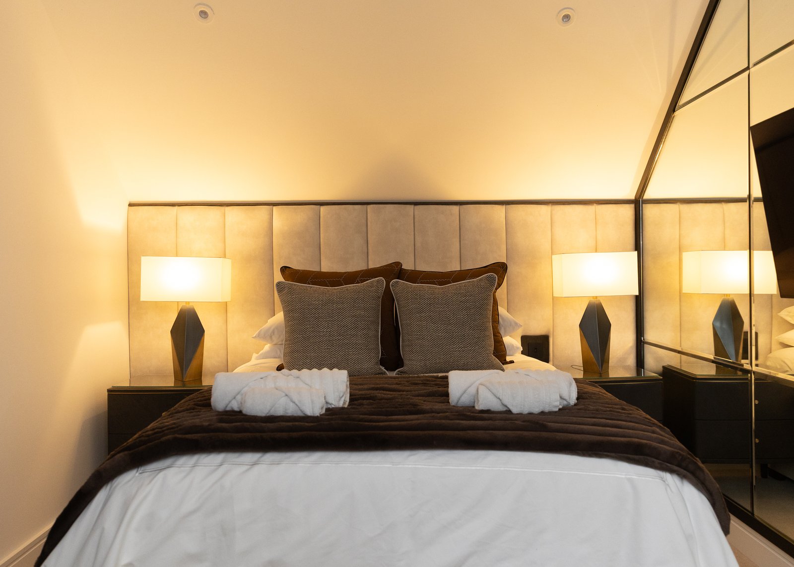 Sycamore second bedroom, a mushroom velvet-panelled headboard running the width of the wall, a pair of faceted bronze lamps with white shades, chocolate bedspread, tweed and rust cushions