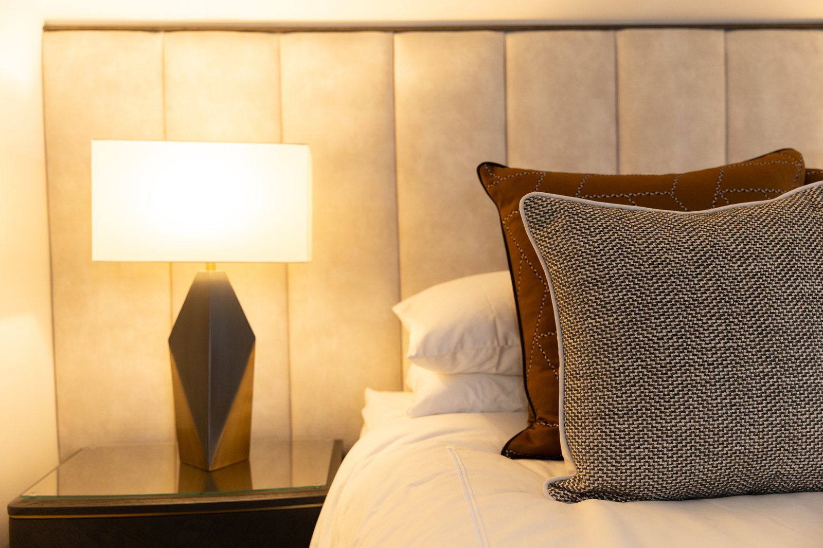 Close detail of the faceted bronze bedside lamp, white shade lit, against the mushroom velvet headboard