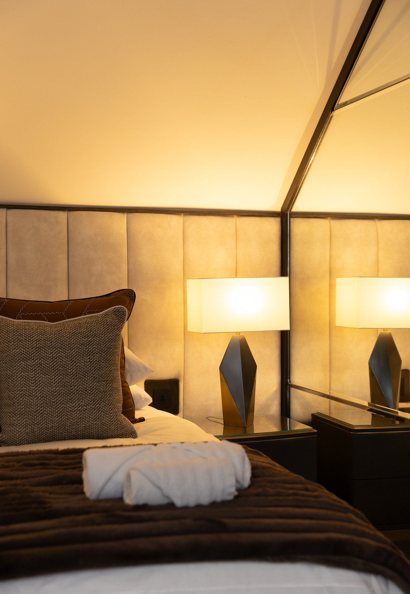 Side view of the second bedroom, mushroom headboard in soft amber light, a folded towel on the bed and the faceted bronze bedside lamp lit