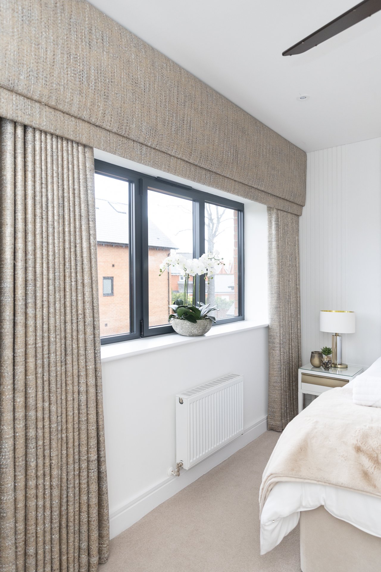 Sycamore third bedroom, a heavy textured linen pelmet and matching curtains framing a window with a white orchid on the sill, mirrored bedside table with a glass lamp, cream bedding