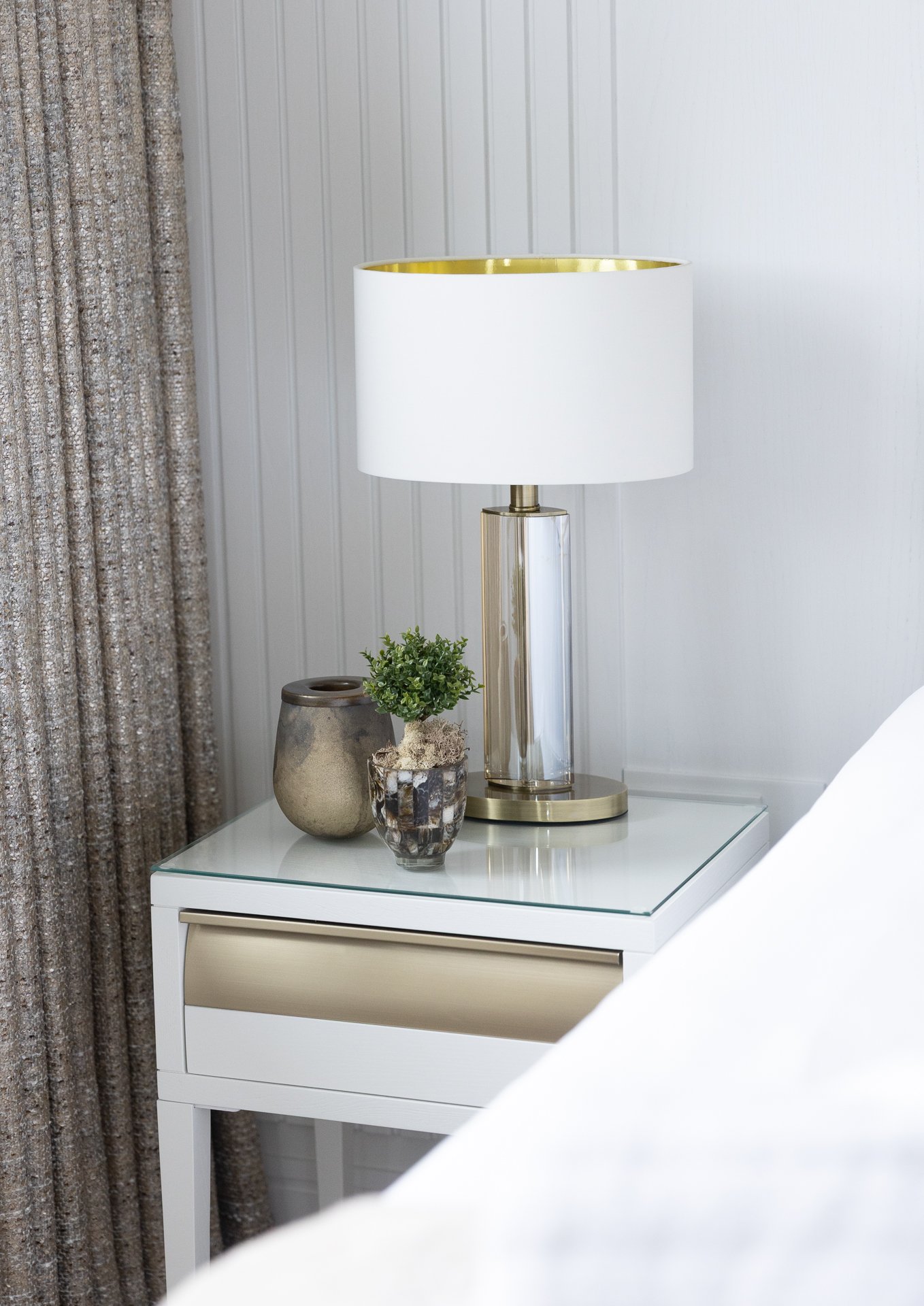 The glass-based bedside lamp with a gold-lined shade, next to a small green plant and a pewter vessel on a white mirrored bedside table