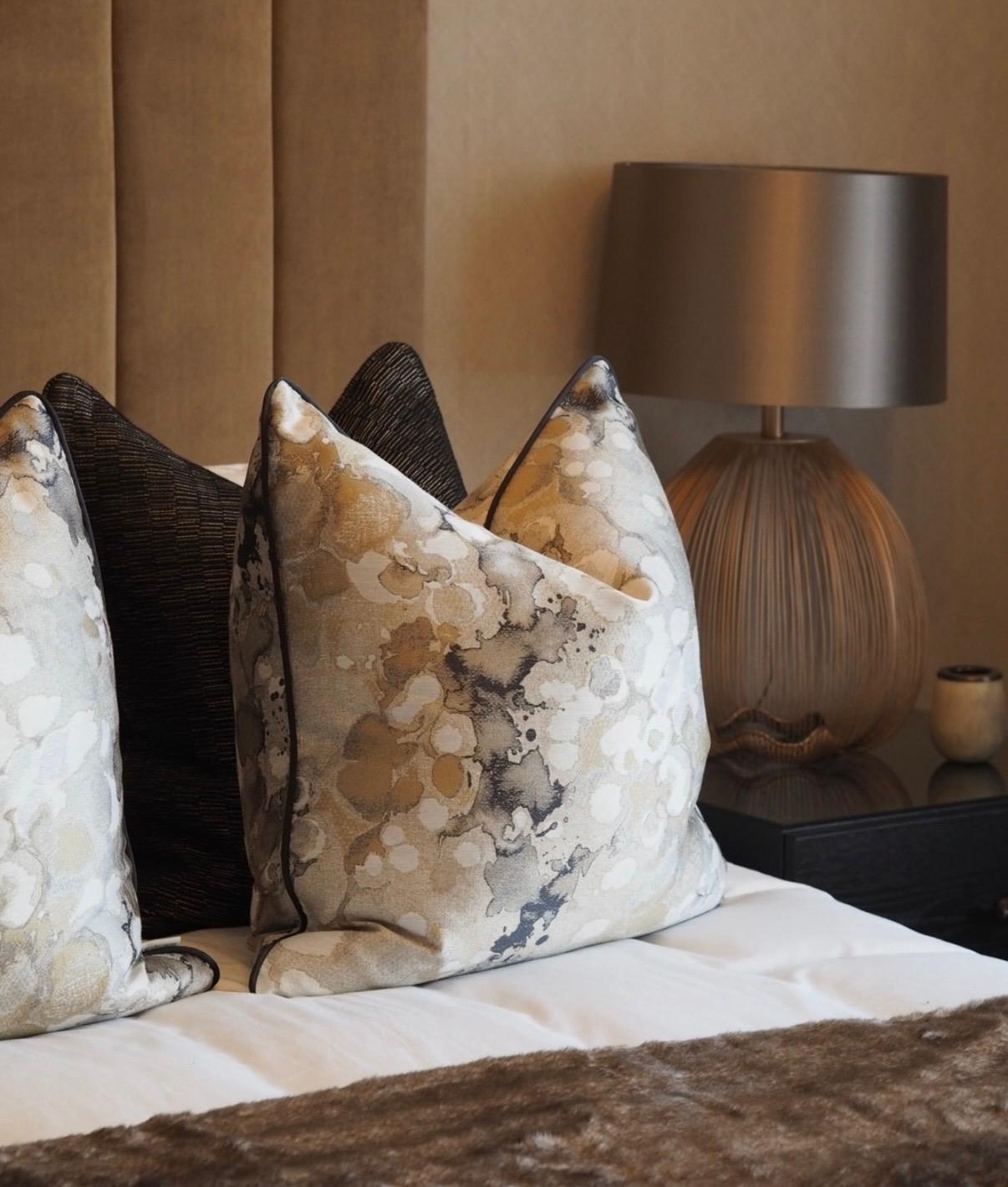 Marbled abstract cushions in cream, ochre and grey beside a dark woven cushion, against a caramel-toned headboard with a ribbed copper lamp — Designers Guild colour-and-pattern handwriting