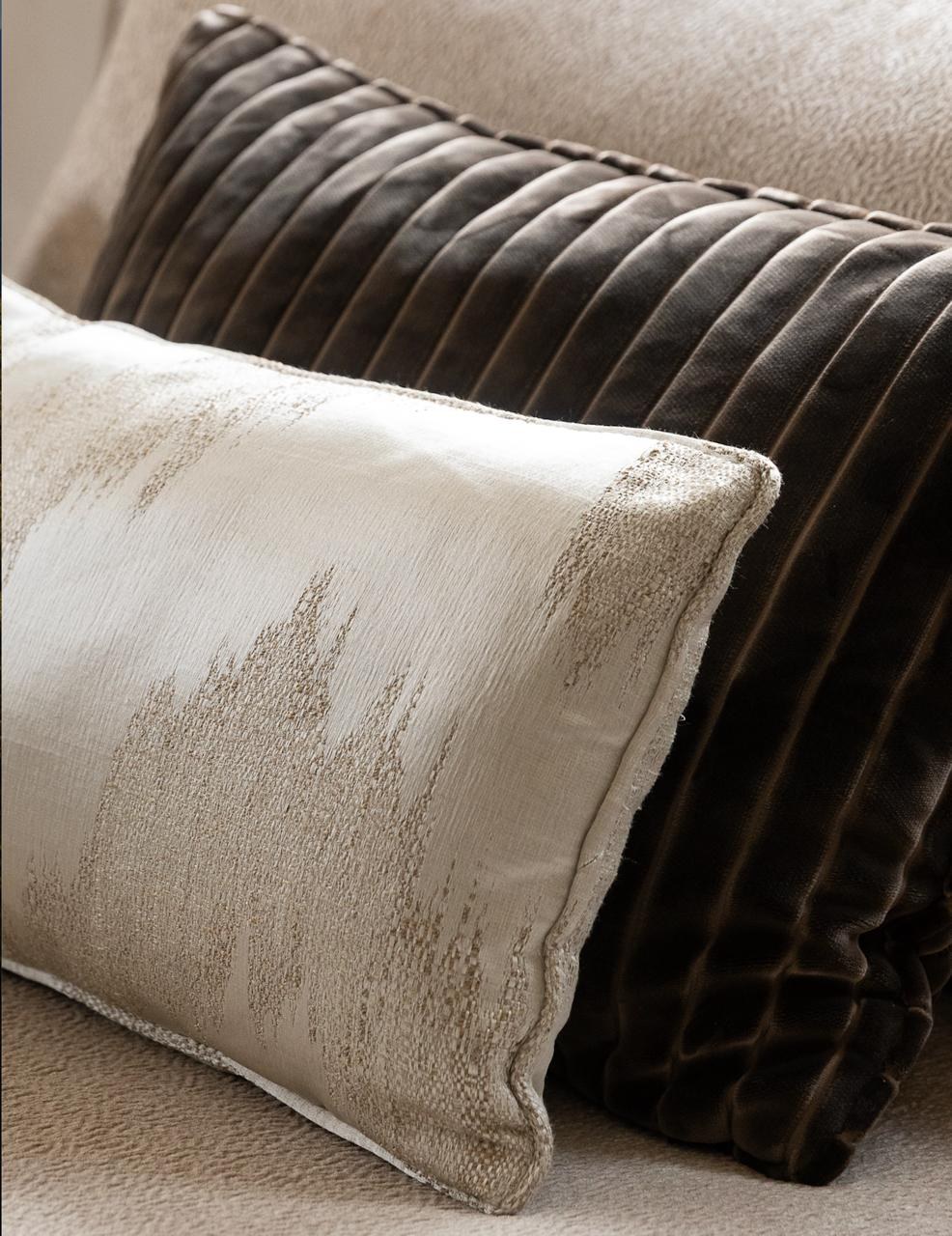A cream linen cushion with a subtle ikat motif beside a dark brown pleated velvet bolster, in the soft Zinc Textile palette